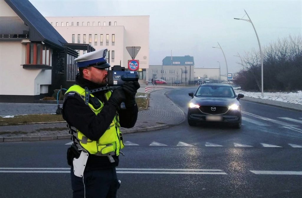 policjant ruchu drogowego z laserowym miernikiem predkosci w tle jadacy samochod