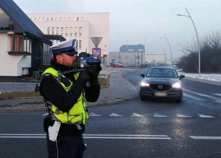 policjant ruchu drogowego z laserowym miernikiem predkosci w tle jadacy samochod