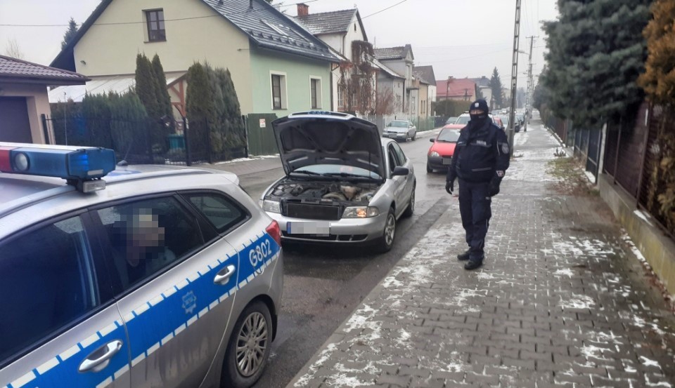 srebrny samochod z otwarta maska przy nim umundurowany policjant obok oznakowany radiowoz