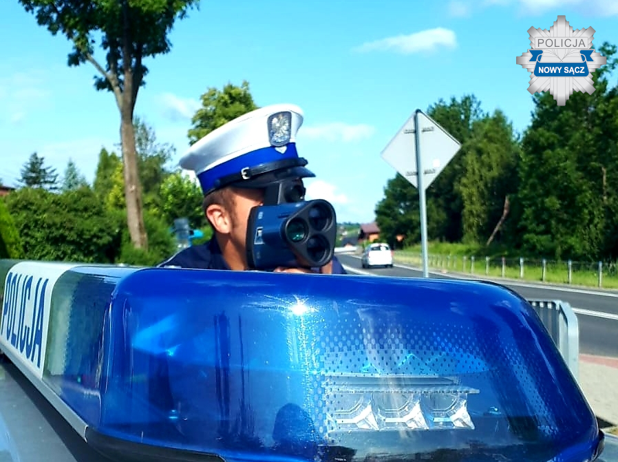 policjant z laserowym miernikiem predkosci obok niebieska lampa na dachu radiowozu z napisem POLICJA poziom