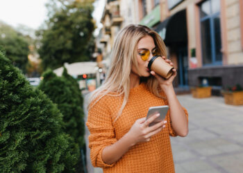Enchanting blonde young woman waiting for phone message while drinking coffee on the street. Stylish girl in yellow sweater posing on alley holding smartphone and enjoying cappuccino.