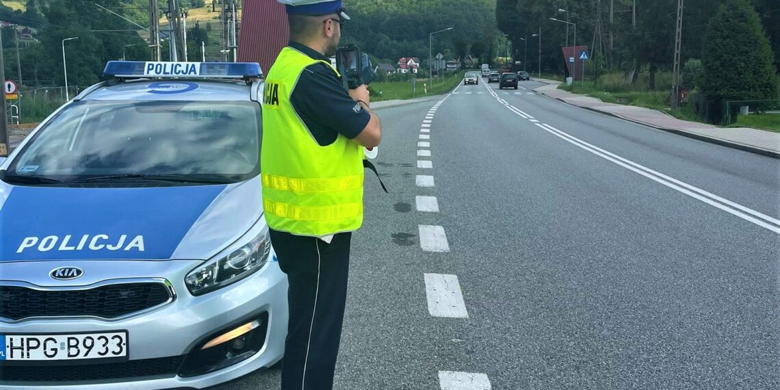 umundurowany policjant drogowki z laserowym miernikiem predkosci obok radiowoz w oddali jadace samochody