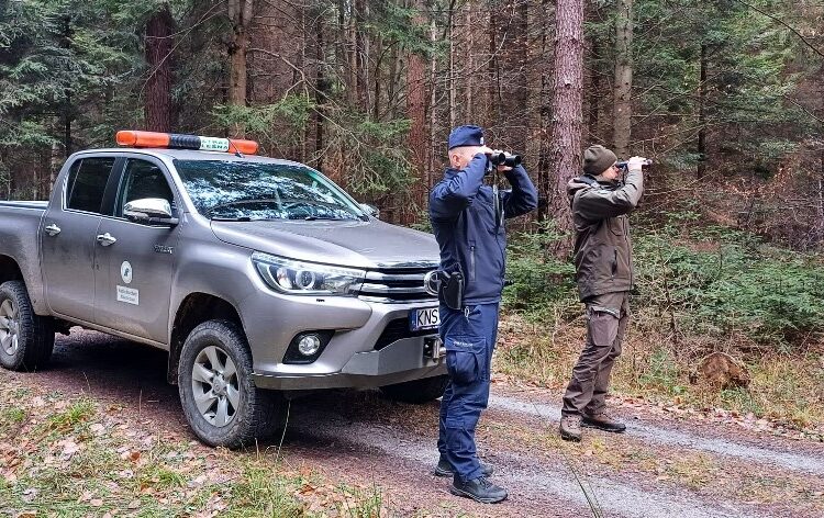 policjant i straznik lesny patrza przez lornetki obok pojazd sluzbowy Strazy Lesnej