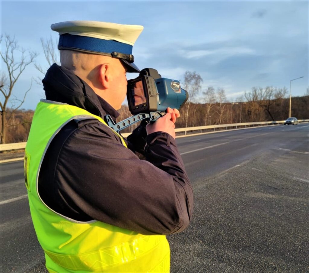 policjant ruchu drogowego z laserowym miernikiem predkosci w oddali jadacy samochod