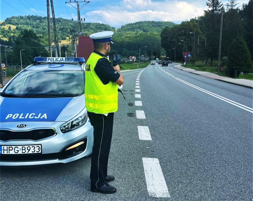 umundurowany policjant drogowki z laserowym miernikiem predkosci obok radiowoz w oddali jadace samochody